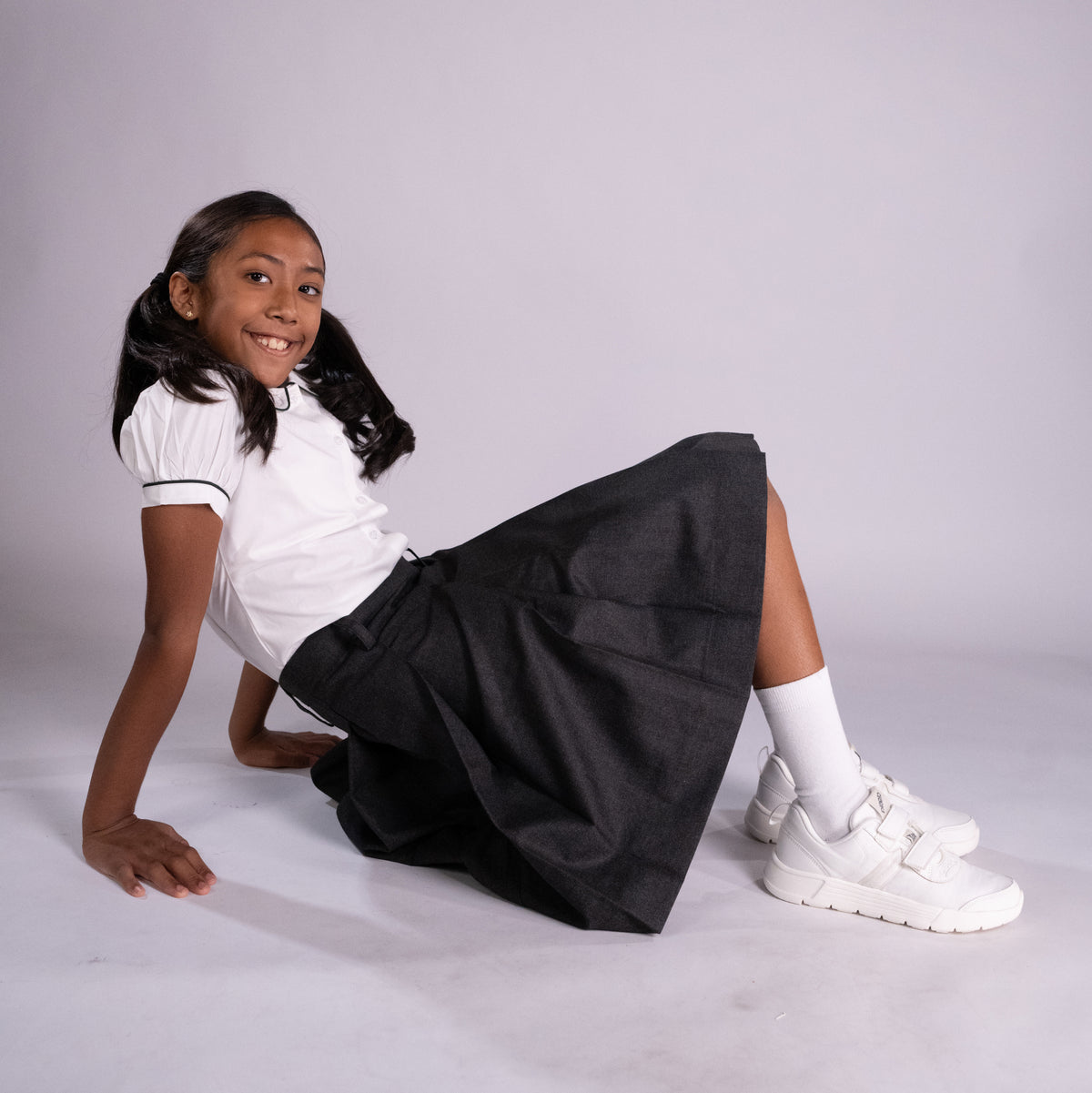 Young girl sitting on a white surface wearing a white shirt, black skirt, and white sneakers.
